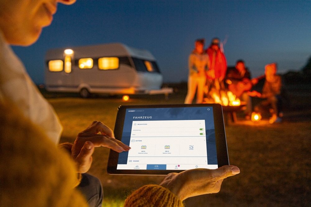 Person hält Tablet mit Fahrzeug-App beim Camping am Lagerfeuer