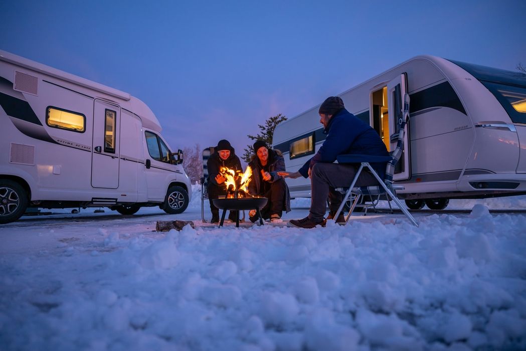 Familie sitzt am Lagerfeuer zwischen zwei Wohnmobilen im Schnee bei Dämmerung