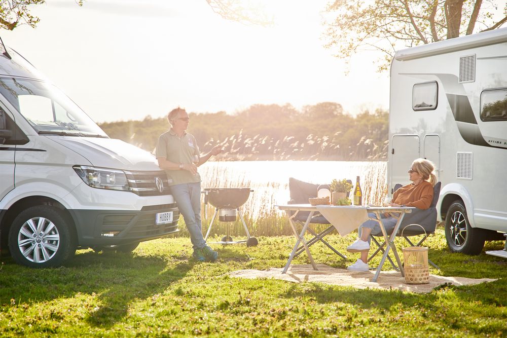 Zwei Personen beim Camping am See mit Wohnmobilen und Campingausrüstung bei Sonnenuntergang