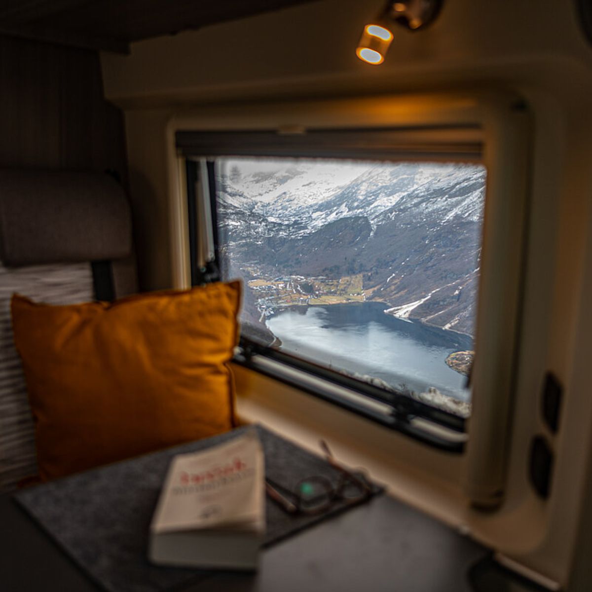 Ausblick aus Wohnwagenfenster auf schneebedeckte Berge und See mit orangem Kissen