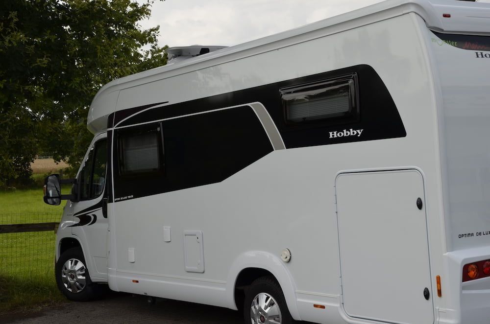 Weißes Hobby-Wohnmobil mit Fensterblenden steht auf Wiese neben Bäumen