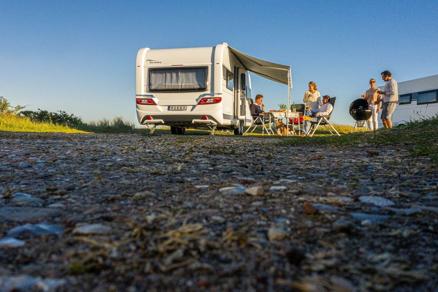 Familie sitzt vor einem Hobby-Wohnwagen mit ausgefahrenem Vorzelt am Campingplatz